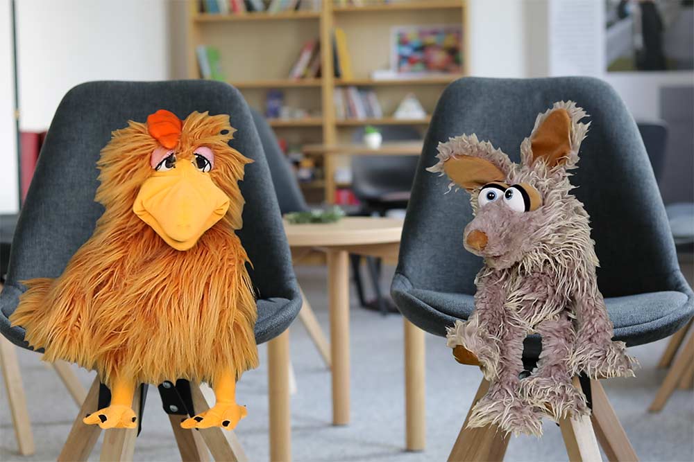 Die Puppen Herta (eine Henne) und Flausi (ein Monster) sitzen jeweils auf einem Stuhl und unterhalten sich.