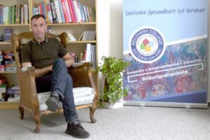 Ein Mann sitzt im Recovery College Gütersloh-OWL auf einem alten Sessel. Im Hintergrund ein Bücherregal und Rollup vom Recovery College Gütersloh-OWL.