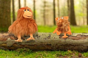 Die Puppen Herta (eine Henne) und Flausi (ein Monster) sitzen im Wald auf einem Baumstamm