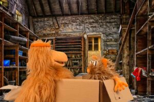 Die Puppen Herta (eine Henne) und Flausi (ein Monster) befinden sich in einem alten, schmuddeligen Lagerraum. Flausi sitzt in einem Pappkarton und Herta steht neben dem Pappkarton.