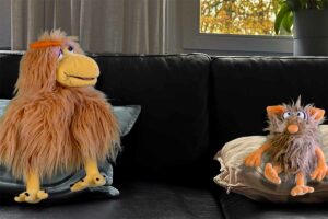 Die Puppen Herta (eine Henne) und Flausi (ein Monster) sitzen auf einem Sofa und unterhalten sich.