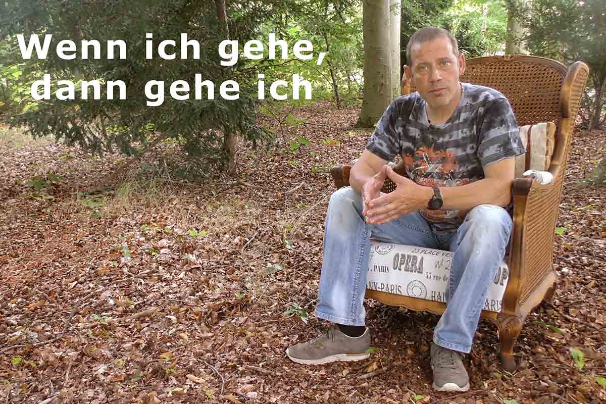 Ein Mann sitzt in einem kleinen Waldstück mit Laub auf dem Boden auf einem alten Sessel.
